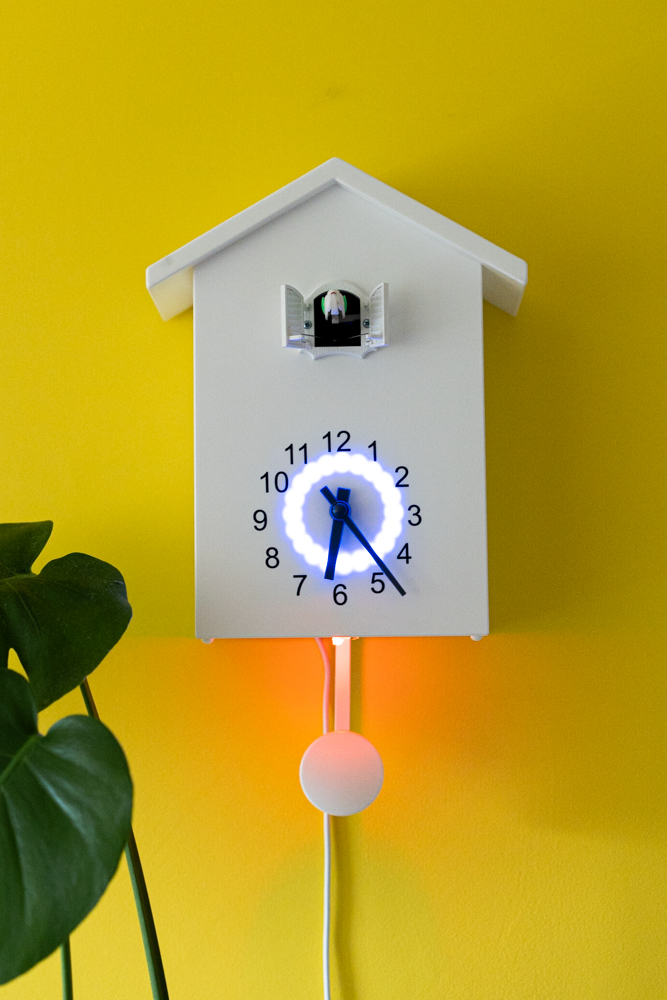 While cuckoo clock on a yellow wall. The clock has a ring of light around the dial and a red light shining onto the pendulum. 